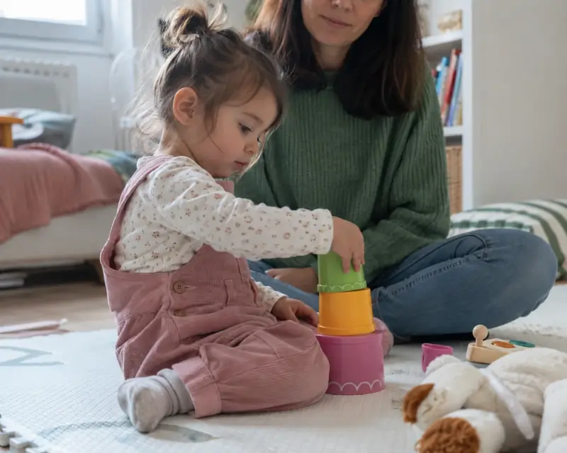 Una niña pequeña apila vasos de colores mientras una adulta la acompaña en el suelo.