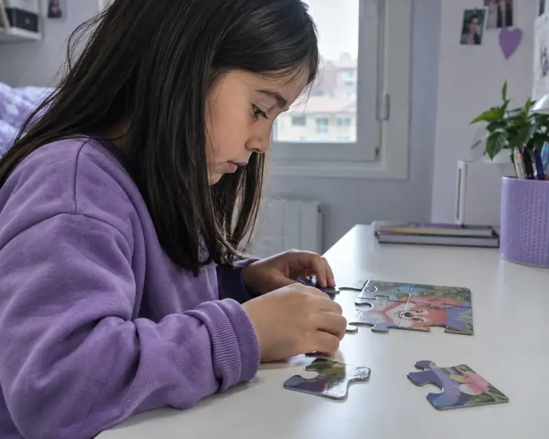 Una niña con sudadera morada se concentra en completar un puzle sobre una mesa.