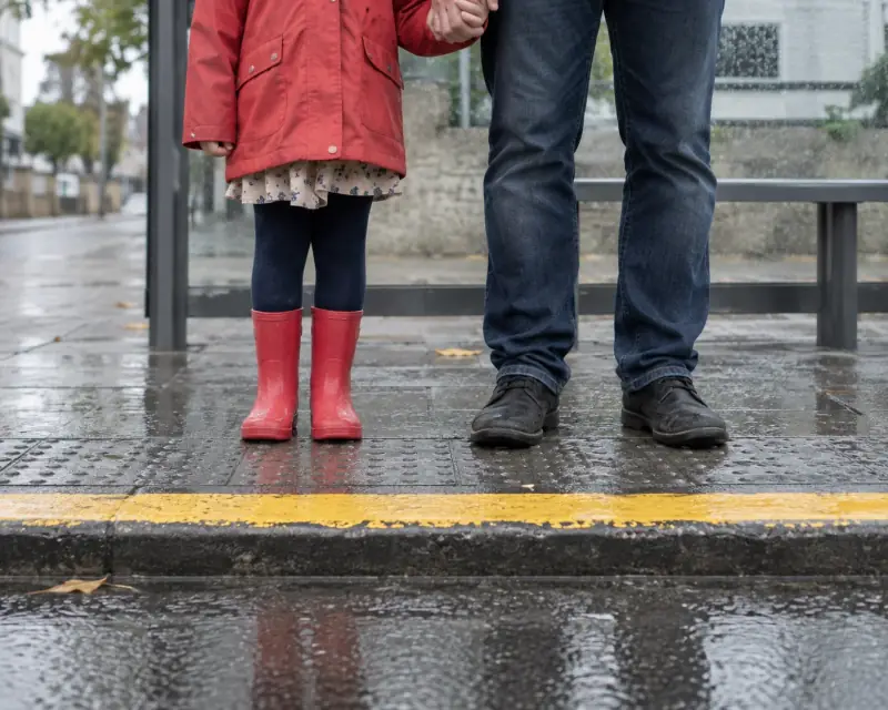 Una niña con botas rojas espera junto a un adulto en una parada de autobús.