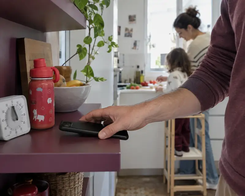 Una mano deja el móvil en una estantería mientras una adulta y una niña cocinan al fondo.