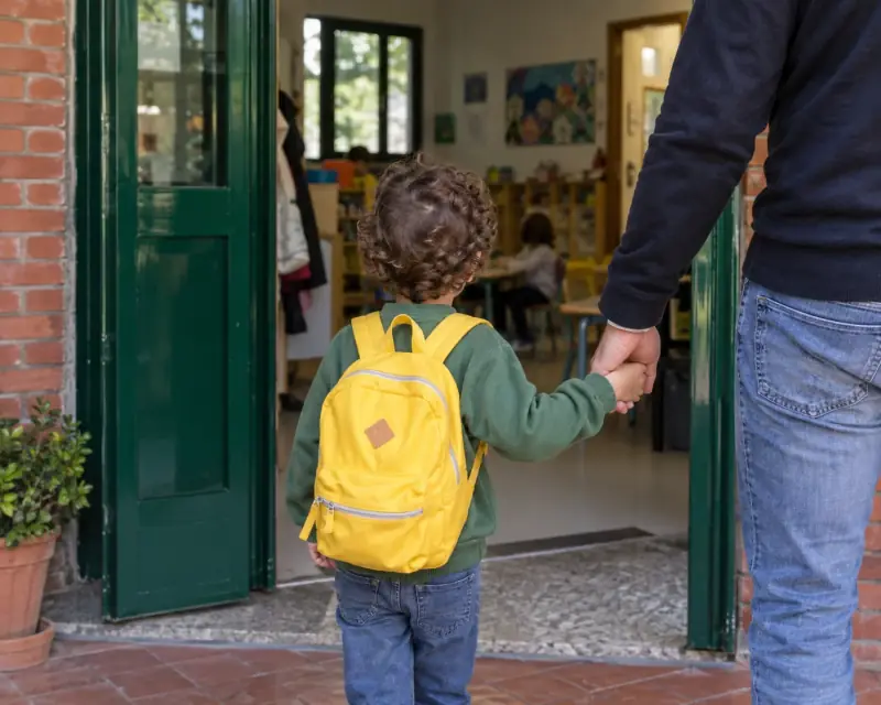 Un niño con mochila amarilla entra en el colegio de la mano de un adulto.