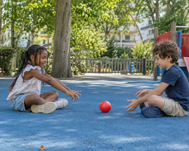 Dos niños sentados frente a frente se pasan una pelota roja en un parque.
