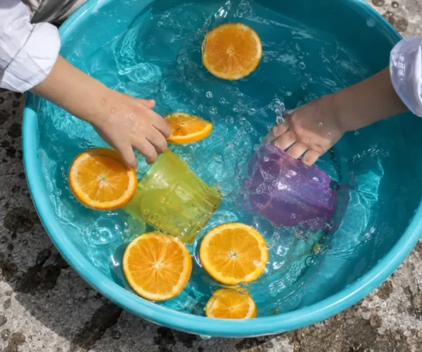 Manos infantiles juegan con vasos y rodajas de naranja en un recipiente con agua.